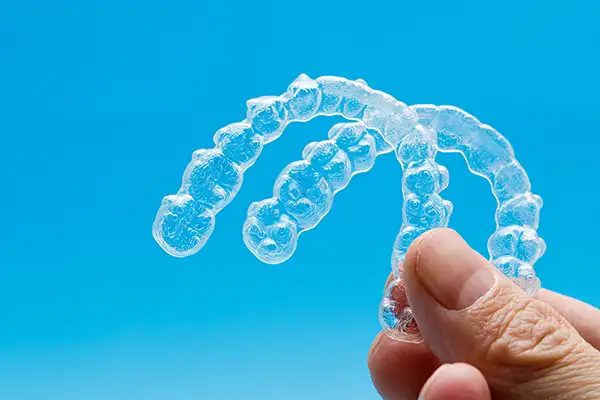 Close-up of a hand holding upper and lower Invisalign clear aligner trays, the blue background shining through to showcase the trays' transparent, light material.